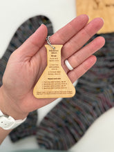 Load image into Gallery viewer, Hand holding a wooden keychain with text against a blurred background of colorful fabric.